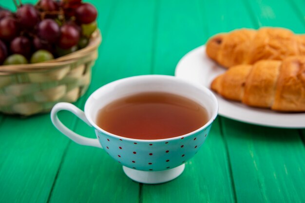 Vista lateral del desayuno con croissants en un plato, taza de té y una canasta de endrinas de uva sobre fondo verde