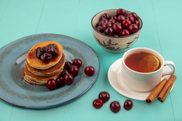 Vista lateral del desayuno con una taza de té y canela en un platillo y panqueques con cerezas en un plato y un tazón de cerezas sobre fondo azul.