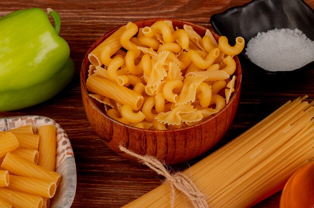 Vista lateral de diferentes macarrones en un tazón y en un plato con sal de pimienta en la mesa de madera