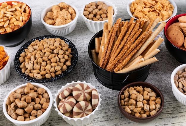 Vista lateral de diferentes tipos de bocadillos como nueces, galletas y galletas en tazones sobre una superficie de madera blanca horizontal