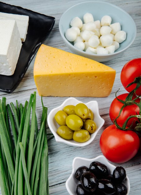 Vista lateral de diferentes tipos de queso con cebolla verde, aceitunas en vinagre y tomates frescos en la mesa de madera gris