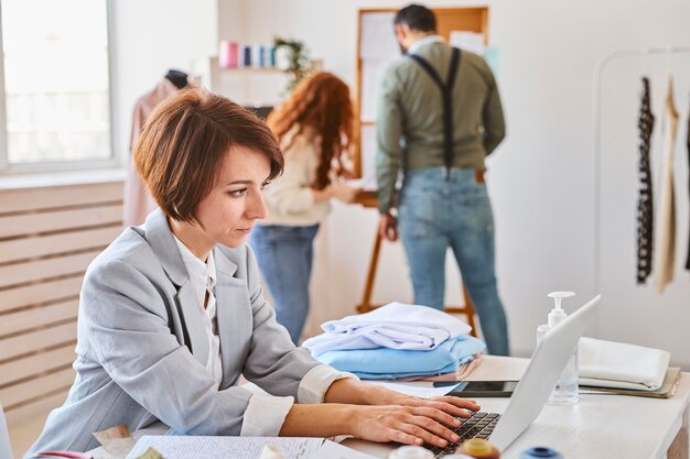 Vista lateral de la diseñadora de moda femenina que trabaja en el atelier con una computadora portátil y colegas