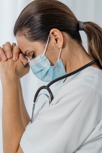 Vista lateral de la doctora con máscara médica y estetoscopio rezando