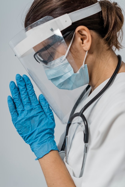 Vista lateral de la doctora con máscara médica y protector facial rezando