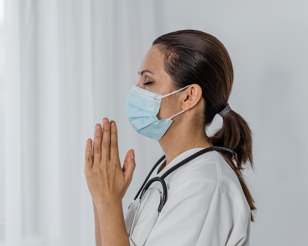 Vista lateral de la doctora con máscara médica rezando