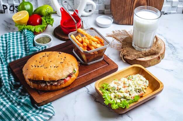 Vista lateral de doner kebab con carne en pan de pita sobre una tabla de madera con ensalada de verduras mixtas papas fritas y bebida ayran