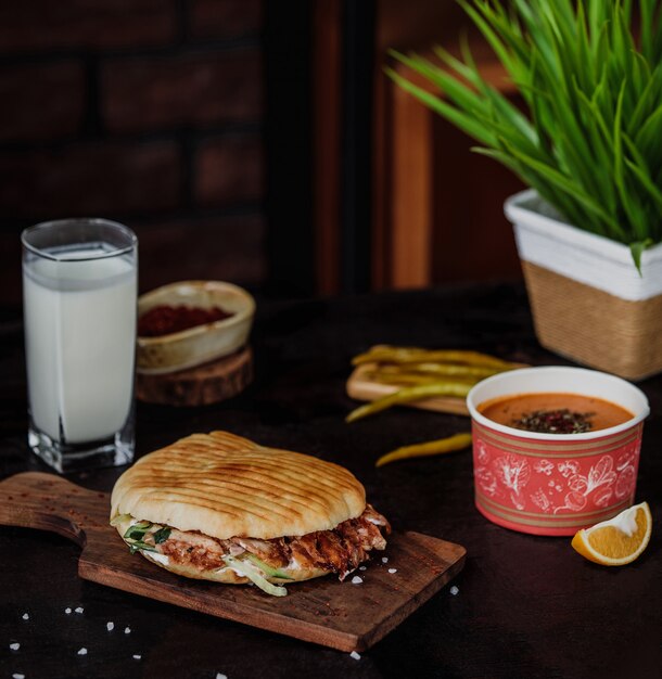 Vista lateral de doner kebab en pan de pita sobre una tabla de madera servida con sopa de dintel, bebida ayran y pimientos verdes encurtidos en la pared de madera