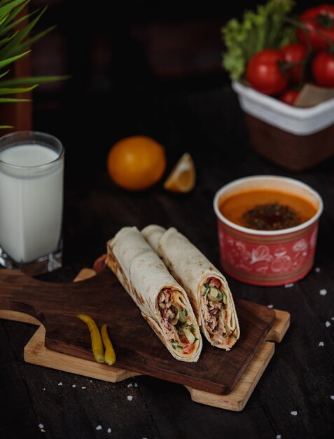 Vista lateral de doner de pollo envuelto en lavash sobre una tabla de madera servida con sopa de dintel y bebida ayran sobre la mesa