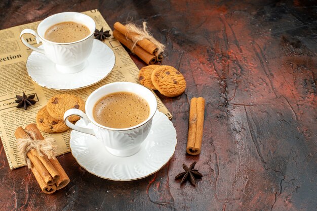 Vista lateral de dos tazas de café, galletas, canela, limas en un periódico viejo sobre una superficie oscura.