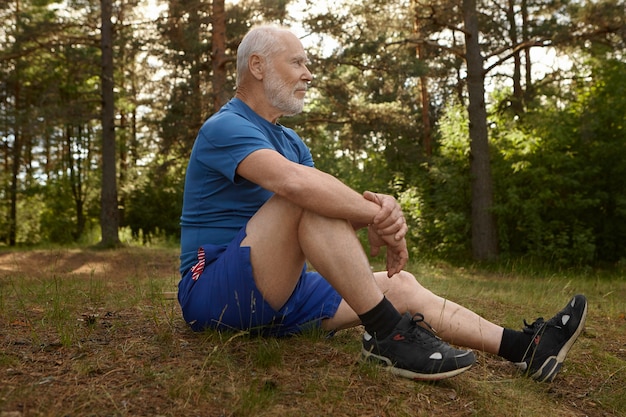 Vista lateral del elegante jubilado masculino con barba contemplando un bonito paisaje sentado en el borde del bosque, relajándose después del entrenamiento cardiovascular matutino, abrazando la rodilla con ambos brazos, con mirada tranquila y pacífica