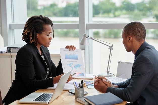 Vista lateral de la empresaria mostrando gráficos analíticos a su compañero de trabajo masculino