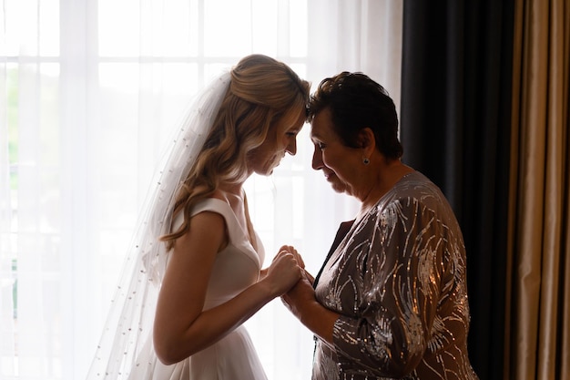 Vista lateral de la encantadora madre y la linda novia que visten trajes de celebración de pie cara a cara cogidos de la mano y sintiéndose felices durante el día de la boda xA