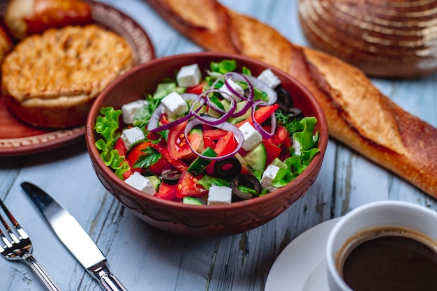 Vista lateral ensalada griega con queso blanco tomate cebolla roja lechuga pepino aceituna negra y taza de café sobre la mesa