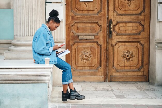 Vista lateral de una estudiante afroamericana casual con chaqueta vaquera con teléfono celular y libro estudiando cuidadosamente en la calle de la ciudad