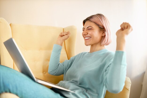 Vista lateral de la feliz mujer joven emocional independiente en ropa casual sentada en un sillón con una computadora portátil en su regazo, apretando los puños, emocionada por la gran oferta de trabajo, exclamando