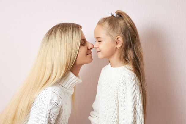 Vista lateral de la feliz mujer joven con largo cabello rubio que va a besar a su encantadora hijita posando con las puntas de las narices presionadas entre sí. Amor, familia, generaciones y relaciones