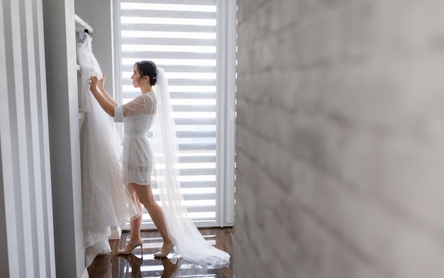 Vista lateral de la feliz novia con velo largo que se está preparando para el día de la boda en la habitación, vistiéndose con un vestido de novia