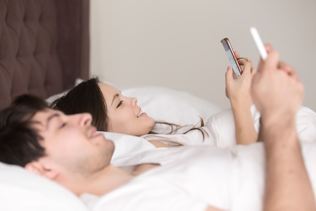 Vista lateral de la feliz pareja joven usando teléfonos inteligentes en la cama