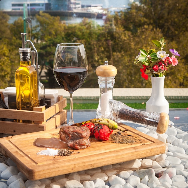 Vista lateral de filete de carne con pomato sobre tabla de madera con copa de vino en la mesa de cristal con vista al mar