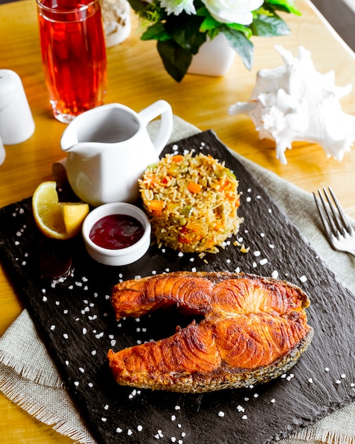 Vista lateral de filete de pescado rojo frito con arroz con verduras una rodaja de limón y salsa de granada en el tablero