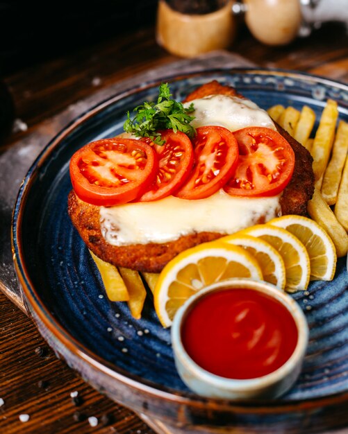 Vista lateral del filete de pollo empanizado frito con queso servido con rodajas de tomate, limones, salsa de tomate y papas fritas en rústica