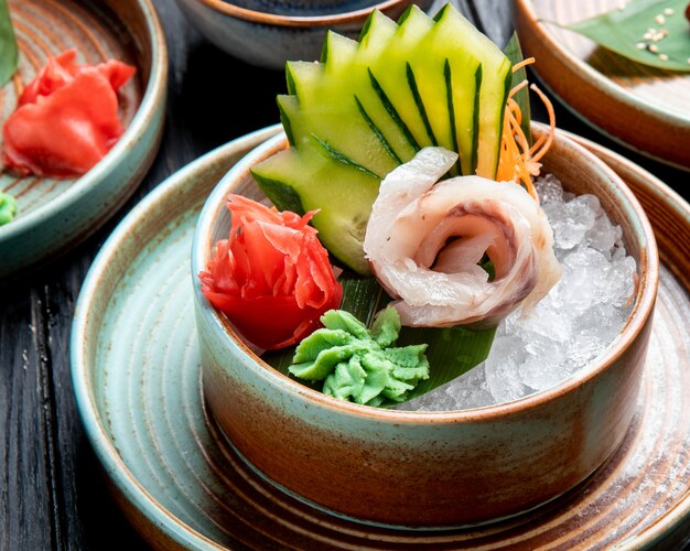 Vista lateral de filetes de arenque marinados con pepinos en rodajas de jengibre y salsa de wasabi en cubitos de hielo en un plato sobre la mesa