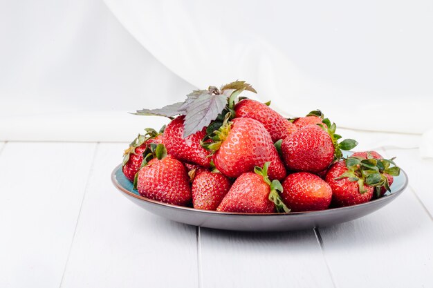 Vista lateral fresa fresca en un plato con albahaca sobre fondo blanco de madera