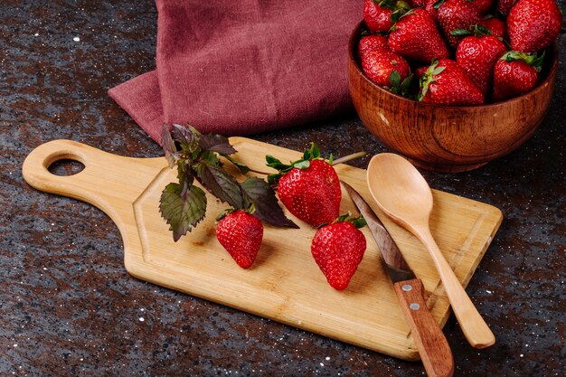 Vista lateral de fresa fresca en un tazón de albahaca cuchillo cuchara y tablero sobre fondo negro