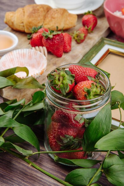 Vista lateral de fresas frescas maduras en un frasco de vidrio y hojas verdes croissant en madera rústica
