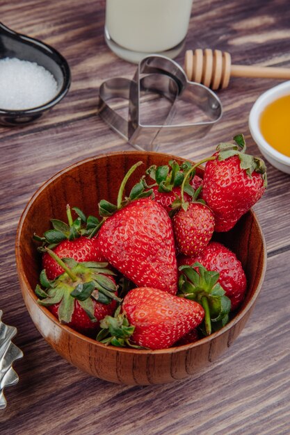 Vista lateral de fresas frescas maduras en un tazón de madera con cortadores de galletas y miel en rústico