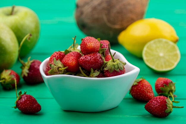 Vista lateral de fresas rojas frescas en un recipiente blanco con limón, lima, manzana sobre un fondo verde