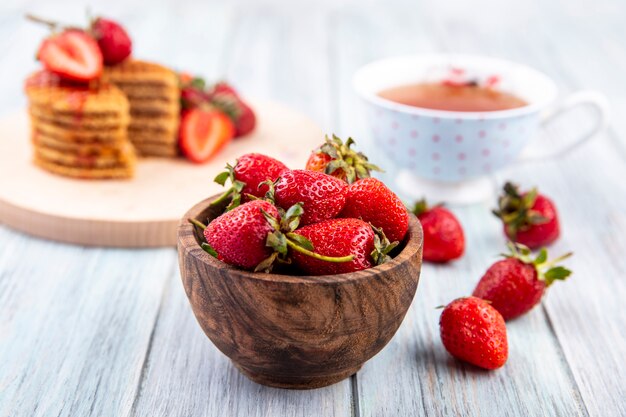 Vista lateral de fresas en un tazón con té y galletas waffle sobre madera