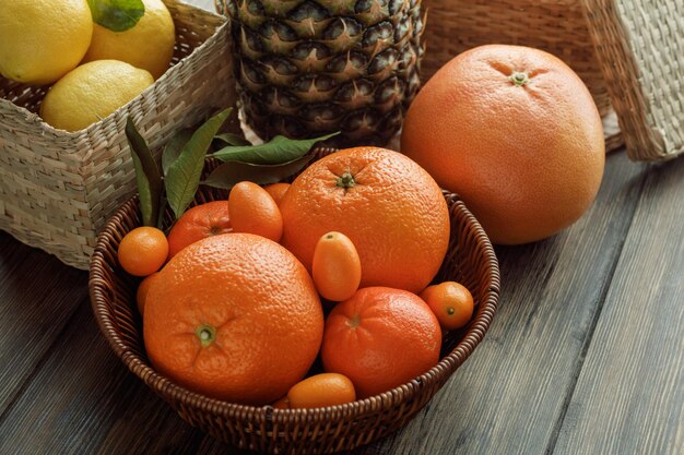 Vista lateral de frutas cítricas como naranja mandarina kumquat en canasta con canasta de limones piña sobre fondo de madera