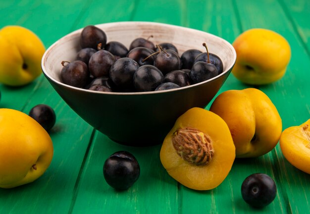 Vista lateral de frutas como endrinas en un tazón y albaricoques con medio corte sobre fondo verde