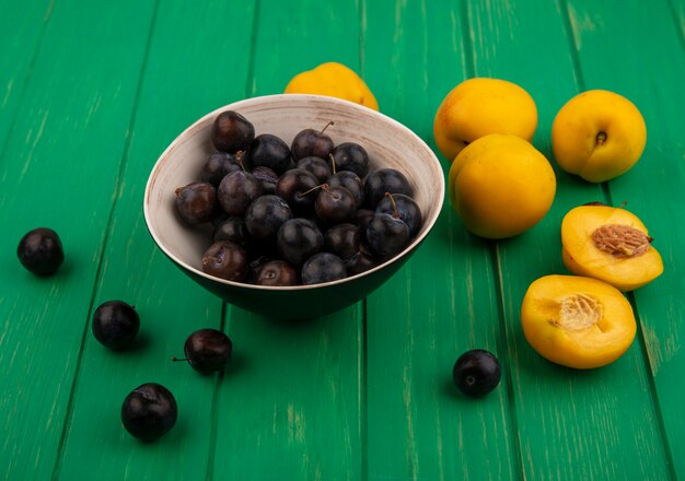 Vista lateral de frutas como endrinas en un tazón y albaricoques con medio corte sobre fondo verde