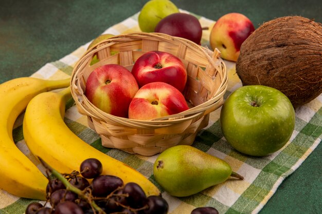 Vista lateral de frutas como melocotón en canasta y coco plátano pera uva en tela escocesa sobre fondo verde