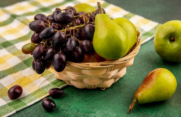 Vista lateral de frutas como melocotón uva y pera en canasta sobre tela escocesa con manzana sobre fondo verde