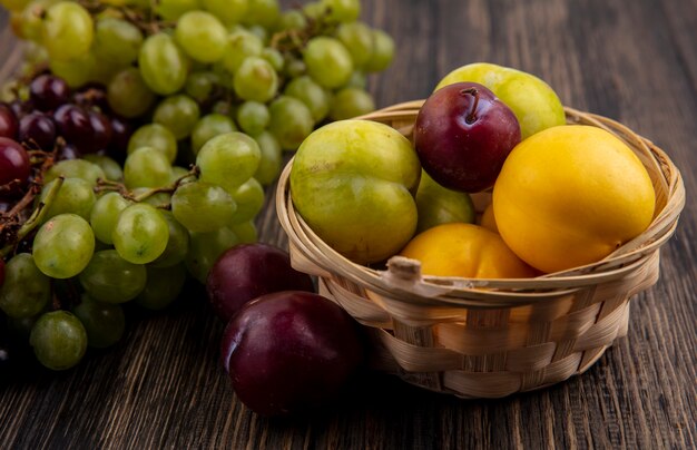 Vista lateral de frutas como nectacots verde y sabor king pluots en canasta con uvas sobre fondo de madera