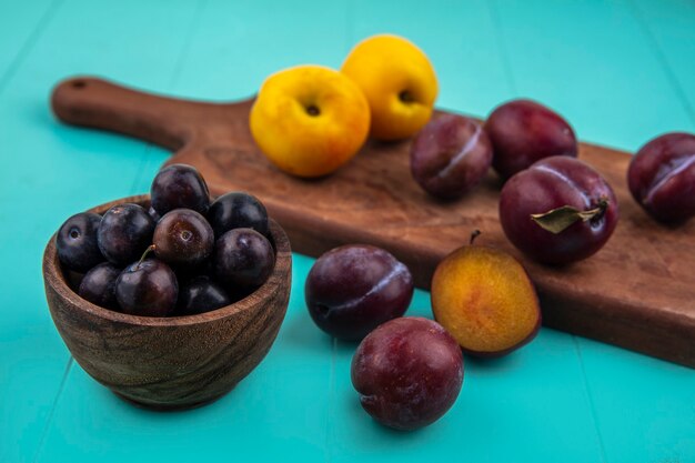 Vista lateral de frutas como tazón de bayas de uva con nectacots y pluots sobre tabla de cortar sobre fondo azul.