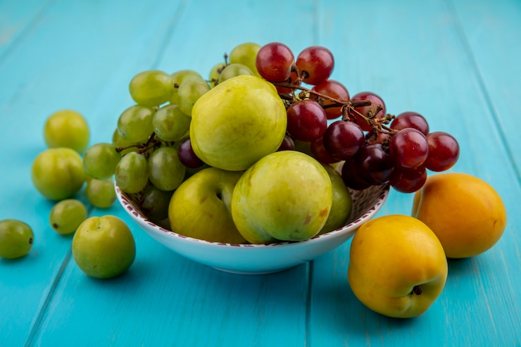 Foto gratuito vista lateral de frutas como uvas pluots verdes en un tazón y patrón de ciruelas y nectacots sobre fondo azul.
