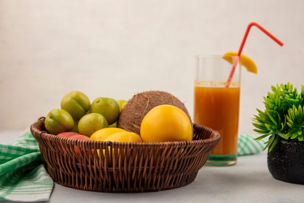 Vista lateral de frutas frescas como ciruelas cereza verde coco melocotones amarillos en un cubo con jugo de durazno fresco en un vaso sobre un fondo blanco