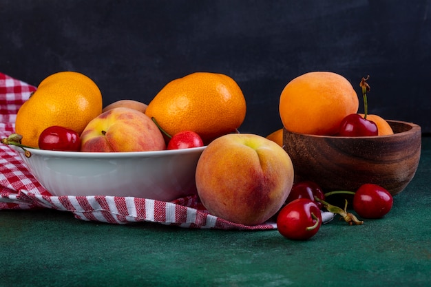 Vista lateral de frutas maduras frescas, mandarinas, duraznos y cerezas rojas en un tazón sobre tela escocesa oscura