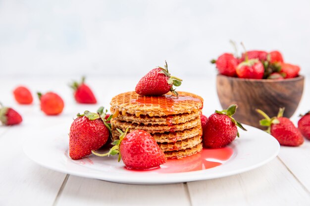 Vista lateral de galletas waffle con fresas en placa y en tazón y sobre madera