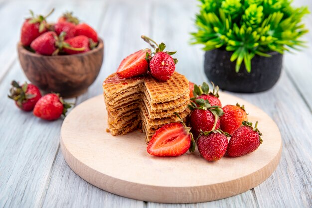 Vista lateral de galletas waffle sobre tabla de cortar y fresas en un tazón y sobre madera