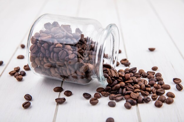 Vista lateral de los granos de café tostados oscuros cayendo de un frasco de vidrio sobre un fondo de madera blanca