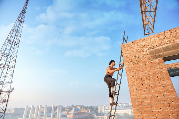 Vista lateral del guapo constructor con el torso desnudo en el sombrero subir la escalera. Escalera apoyada en la pared de ladrillo en un edificio terminado. Alta torre de televisión y paisaje urbano en el fondo.