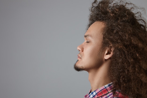 Vista lateral del guapo joven concentrado de raza mixta sin afeitar con voluminoso cabello ondulado cerrando los ojos