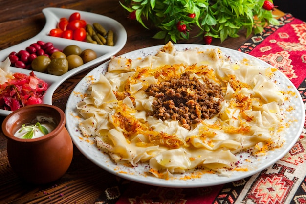 Vista lateral gurú azerbaiyano khingal pasta caucásica con carne picada frita y cebolla con salsa de crema agria y encurtidos sobre un mantel sobre una mesa de madera oscura horizontal