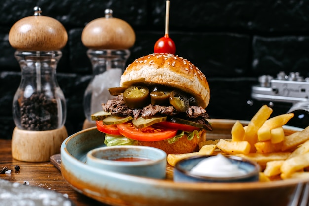 Vista lateral de hamburguesa con encurtidos de carne de res y tomates servidos con papas fritas y salsas en negro