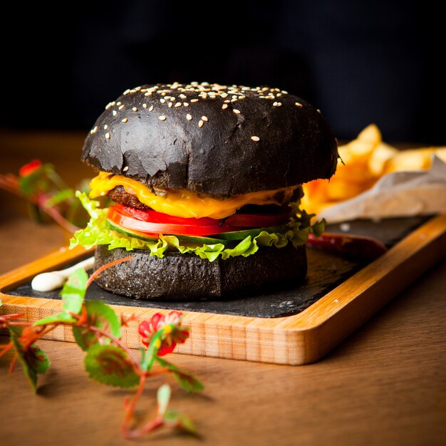 Vista lateral hamburguesa negra con papas fritas y salsa de tomate y flores en bandeja de madera sobre mesa de madera